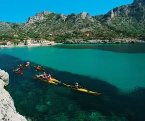 En gruppe kajakroere padler gennem en turkisblå bugt omgivet af klippefyldte bakker. Det klare vand afslører pletter af mørk tang nedenfor. Huse og små bygninger er beliggende ved bunden af klipperne under en klar blå himmel.