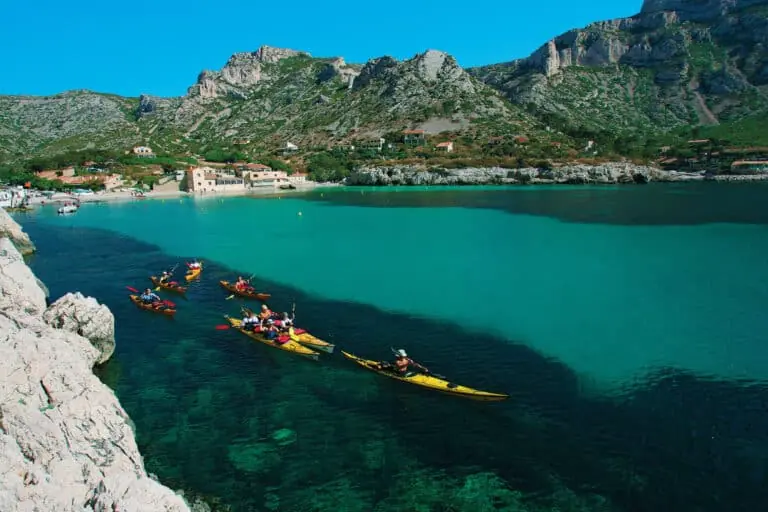 En gruppe kajakroere padler gennem en turkisblå bugt omgivet af klippefyldte bakker. Det klare vand afslører pletter af mørk tang nedenfor. Huse og små bygninger er beliggende ved bunden af klipperne under en klar blå himmel.