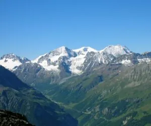 Sneklædte bjerge under en klar blå himmel med frodige grønne dale og stenede skråninger i forgrunden. Barske tinder strækker sig over horisonten og skaber et malerisk alpelandskab.