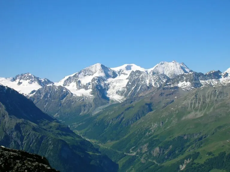 Sneklædte bjerge under en klar blå himmel med frodige grønne dale og stenede skråninger i forgrunden. Barske tinder strækker sig over horisonten og skaber et malerisk alpelandskab.