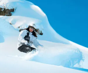 En person klædt i vinterudstyr, inklusive en hvid hjelm og beskyttelsesbriller, snowboards ned ad en sneklædt skråning under en klar blå himmel, og sparker en spray af sne op bag sig. Snedækkede sten er synlige i baggrunden.
