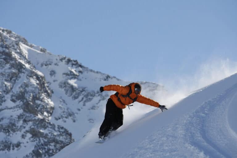 En snowboarder i orange jakke og sorte bukser skærer ned ad en sneklædt bjergskråning mod en klar blå himmel. Sne sprøjter op, mens de laver et skarpt sving, med forrevne, snedækkede toppe i baggrunden.