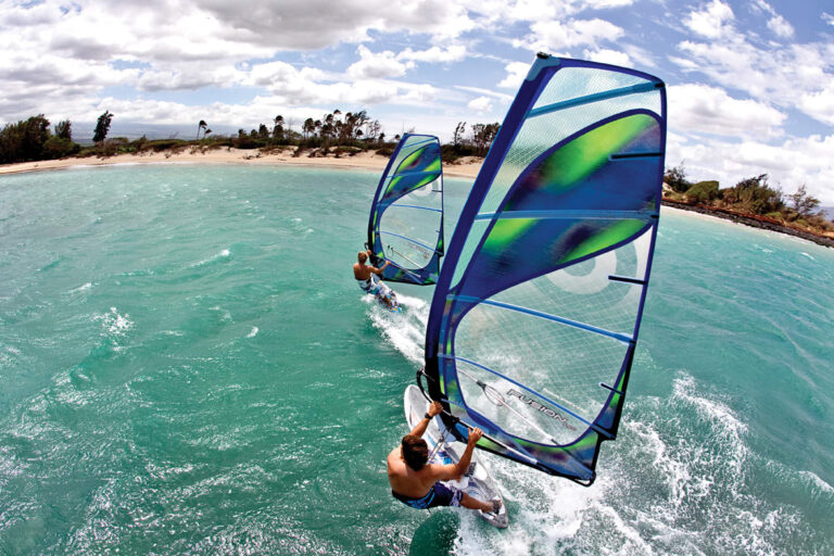 To mennesker windsurfer på et turkisblåt hav nær en sandstrand med palmer. Himlen er delvist overskyet, og windsurferne er iført blå shorts, der hver holder fast i et blåt og grønt sejl.