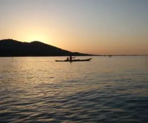 En person med silhuet padler i kano på roligt vand under en solnedgang med et bjergrigt landskab i baggrunden og en varm, glødende himmel.