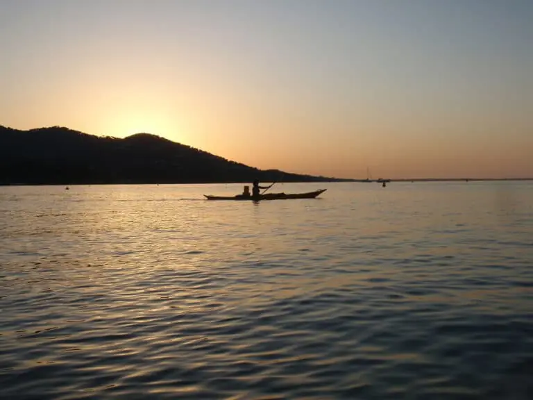En person med silhuet padler i kano på roligt vand under en solnedgang med et bjergrigt landskab i baggrunden og en varm, glødende himmel.