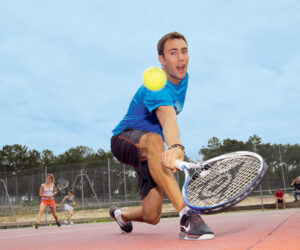 En mand iført en blå skjorte og sorte shorts kaster sig frem på en udendørs tennisbane for at returnere en tennisbold med sin ketsjer. I baggrunden er andre spillere og et hegn synligt under en overskyet himmel.