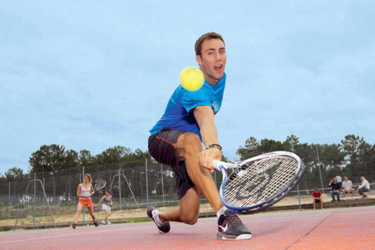 En mand iført en blå skjorte og sorte shorts kaster sig frem på en udendørs tennisbane for at returnere en tennisbold med sin ketsjer. I baggrunden er andre spillere og et hegn synligt under en overskyet himmel.