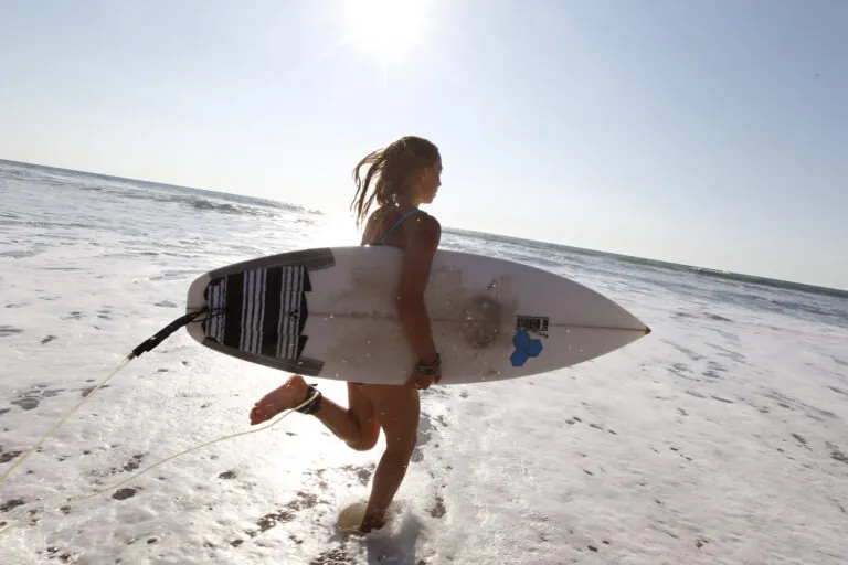 En person med et surfbræt under armen går ud i havet. Solen skinner klart på himlen og kaster refleksioner på vandet. Scenen fanger et øjebliks forventning og spænding før surfing.
