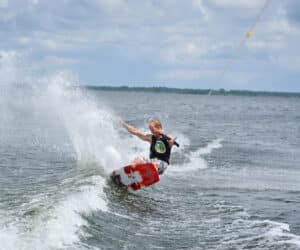 En person, der wakeboarder på en sø, og skaber dygtigt et plask, mens de manøvrerer hen over vandet. Himlen er overskyet, og personen er iført redningsvest og er forbundet til et slæbetov.