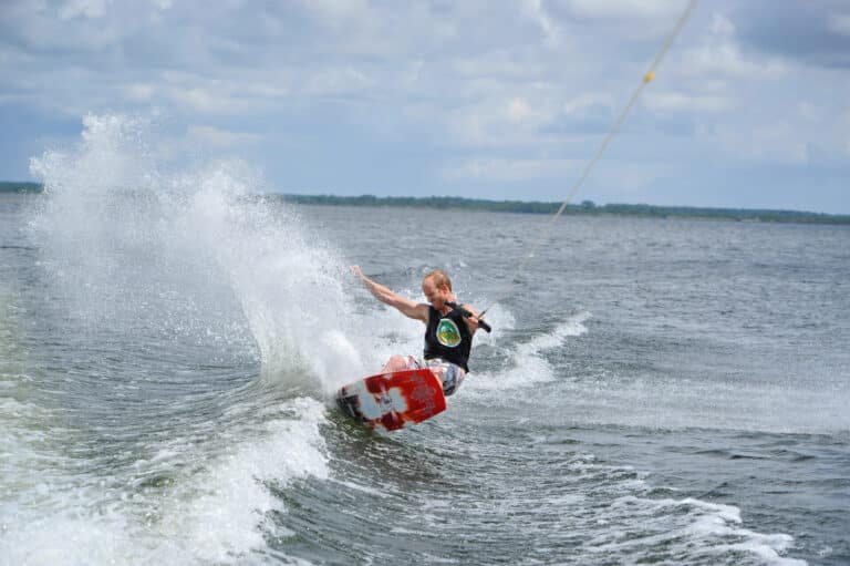 En person, der wakeboarder på en sø, og skaber dygtigt et plask, mens de manøvrerer hen over vandet. Himlen er overskyet, og personen er iført redningsvest og er forbundet til et slæbetov.