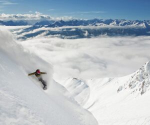 En snowboarder karrer ned ad en stejl, sneklædt bjergskråning med en skyfyldt dal og en fjern bjergkæde under en klar blå himmel.