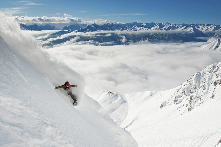 En snowboarder karrer ned ad en stejl, sneklædt bjergskråning med en skyfyldt dal og en fjern bjergkæde under en klar blå himmel.