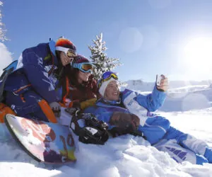 Tre personer i farverigt vinterudstyr sidder sammen i sneen og tager en selfie med en smartphone. Et snowboard er i nærheden, og solen skinner klart på den klare blå himmel og kaster lys over det snedækkede landskab.