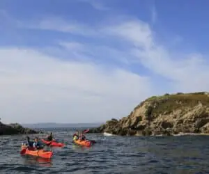 En gruppe kajakroere padler nær en klippeø under en klar blå himmel. Havet er roligt og skaber fredfyldte rammer for det udendørs eventyr.