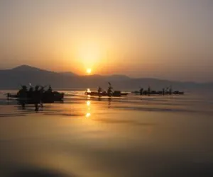 En gruppe kajakroere, der padler på en rolig sø under solopgang. Himlen er en gradient af varme farver, der reflekterer over vandet, med bjerge i baggrunden, der skaber en fredfyldt og fredelig scene.