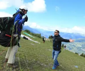 To mænd er på en græsklædt bjergskråning og forbereder sig til paragliding. Den ene er iført sele og hjelm med faldskærm, mens den anden gestikulerer med åbne arme. Baggrunden har en panoramaudsigt over bjerge og dale under en blå himmel med skyer.