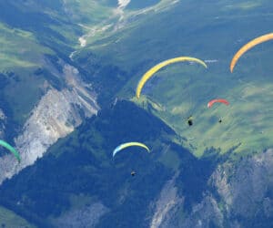 Paraglidere med farverige baldakiner svæver over et frodigt, bjergrigt landskab. De grønne bakker og klippeklipper giver en naturskøn kulisse med fem paragliders synlige under flugten.