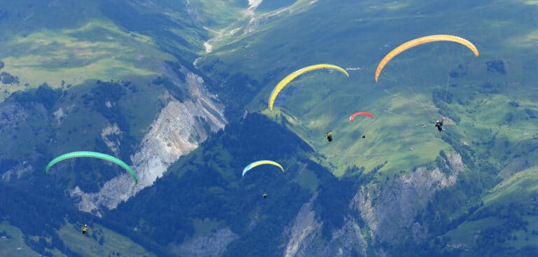 Paraglidere med farverige baldakiner svæver over et frodigt, bjergrigt landskab. De grønne bakker og klippeklipper giver en naturskøn kulisse med fem paragliders synlige under flugten.