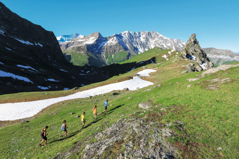 En gruppe på fem personer vandrer op ad en frodig grøn bjergskråning med pletter af sne. Baggrunden viser majestætiske bjerge under en klar blå himmel.