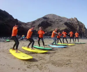 En gruppe mennesker i orange skjorter og sorte våddragter står på surfbrætter opstillet på en sandstrand. De øver sig i at surfe med stenede bakker i baggrunden under en klar himmel.