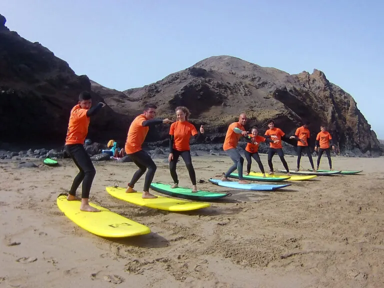En gruppe mennesker i orange skjorter og sorte våddragter står på surfbrætter opstillet på en sandstrand. De øver sig i at surfe med stenede bakker i baggrunden under en klar himmel.