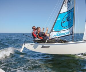 To personer i redningsveste sejler på en lille sejlbåd på åbent vand. Båden vipper til den ene side, mens de navigerer. Himlen er klar, og vandet er roligt.