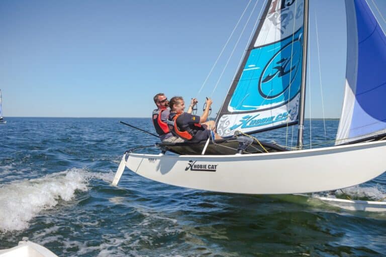 To personer i redningsveste sejler på en lille sejlbåd på åbent vand. Båden vipper til den ene side, mens de navigerer. Himlen er klar, og vandet er roligt.