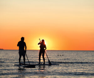 To personer står på paddleboards i havet ved solnedgang. Solen står lavt i horisonten og skaber et varmt orange skær på himlen og reflekterer over vandet. De ser ud til at tale eller holde pause for at nyde udsigten.