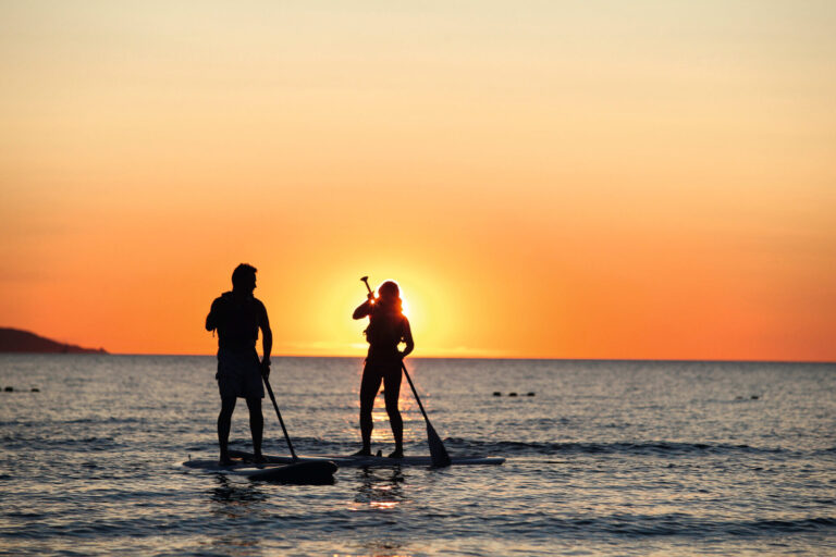 To personer står på paddleboards i havet ved solnedgang. Solen står lavt i horisonten og skaber et varmt orange skær på himlen og reflekterer over vandet. De ser ud til at tale eller holde pause for at nyde udsigten.