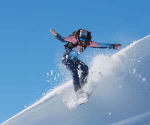 En person i vinterudstyr snowboarder ned ad en sneklædt skråning. De er fanget i luften med frisk pulver bagefter, sat mod en klar blå himmel.