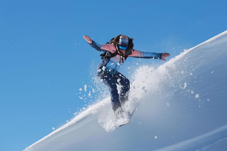 En person i vinterudstyr snowboarder ned ad en sneklædt skråning. De er fanget i luften med frisk pulver bagefter, sat mod en klar blå himmel.