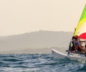 To personer sejler på en farverig Hobie Cat-katamaran i havet. Himlen er klar, og fjerne bakker er synlige i horisonten. En anden sejlbåd ses i baggrunden, hvor bølger forsigtigt bryder rundt om fartøjerne.