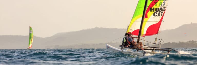 To personer sejler på en farverig Hobie Cat-katamaran i havet. Himlen er klar, og fjerne bakker er synlige i horisonten. En anden sejlbåd ses i baggrunden, hvor bølger forsigtigt bryder rundt om fartøjerne.