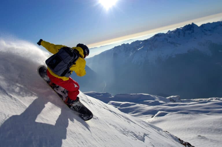 En snowboarder iført en gul jakke og røde bukser går ned ad en snedækket bjergskråning. Solen skinner klart på den blå himmel, med forrevne bjergtoppe synlige i baggrunden. Snesprøjter bag snowboardet.