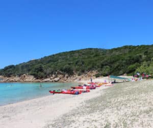 En naturskøn strand med hvidt sand og klart blåt vand. Flere røde kajakker er linet op på kysten. Folk svømmer i havet, og frodige grønne bakker giver en kulisse under en klar blå himmel.