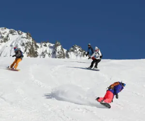 Fire snowboardere kører ned ad en sneklædt bjergskråning under en klar blå himmel. Baggrunden har robuste, snedækkede tinder. Hver snowboarder bærer farverigt vinterudstyr, hvilket skaber en dynamisk scene med udendørs aktiviteter og eventyr.