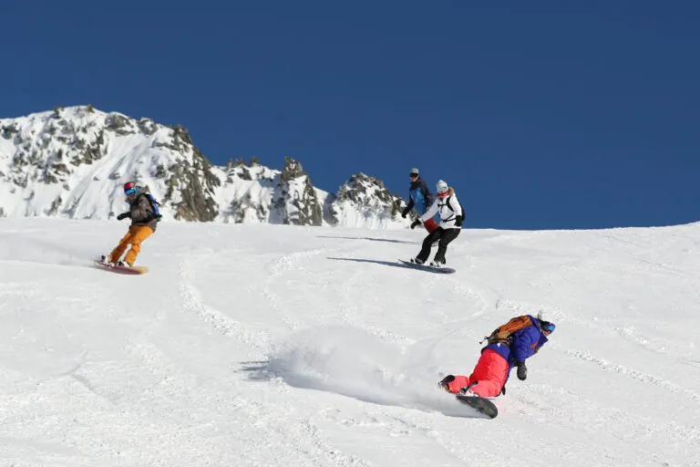 Fire snowboardere kører ned ad en sneklædt bjergskråning under en klar blå himmel. Baggrunden har robuste, snedækkede tinder. Hver snowboarder bærer farverigt vinterudstyr, hvilket skaber en dynamisk scene med udendørs aktiviteter og eventyr.