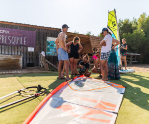 En gruppe mennesker står omkring et windsurfsejl på græsset ved siden af en træbygning med et UCPA-skilt. Nogle er klædt i badetøj. Træer er synlige i baggrunden. Det er en solskinsdag med en person i det fjerne på et paddleboard.