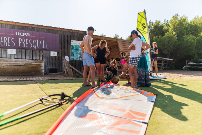 En gruppe mennesker står omkring et windsurfsejl på græsset ved siden af en træbygning med et UCPA-skilt. Nogle er klædt i badetøj. Træer er synlige i baggrunden. Det er en solskinsdag med en person i det fjerne på et paddleboard.