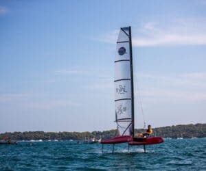 En person sejler en rød hydrofoil sejlbåd på en rolig vandmasse under en klar blå himmel. Andre små både er synlige i det fjerne, nær en trækantet kystlinje.