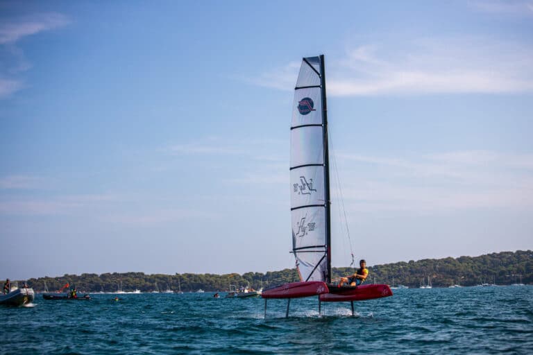 En person sejler en rød hydrofoil sejlbåd på en rolig vandmasse under en klar blå himmel. Andre små både er synlige i det fjerne, nær en trækantet kystlinje.