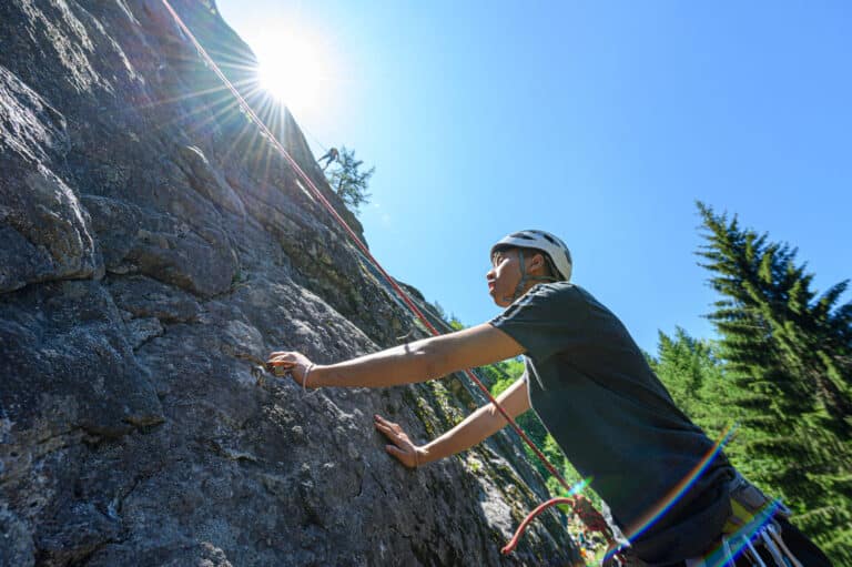 En person iført hjelm og udstyr klatrer op på en klippeflade under en lys, solrig himmel. Sollys skaber en linseudstrålingseffekt. Høje træer er synlige i baggrunden.