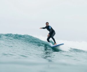 En person i en våddragt surfer på en lille bølge i havet. Det er en overskyet dag, og vandet er roligt. Surferen er balanceret i midten af bølgen, med deres arme strakt for stabilitet.