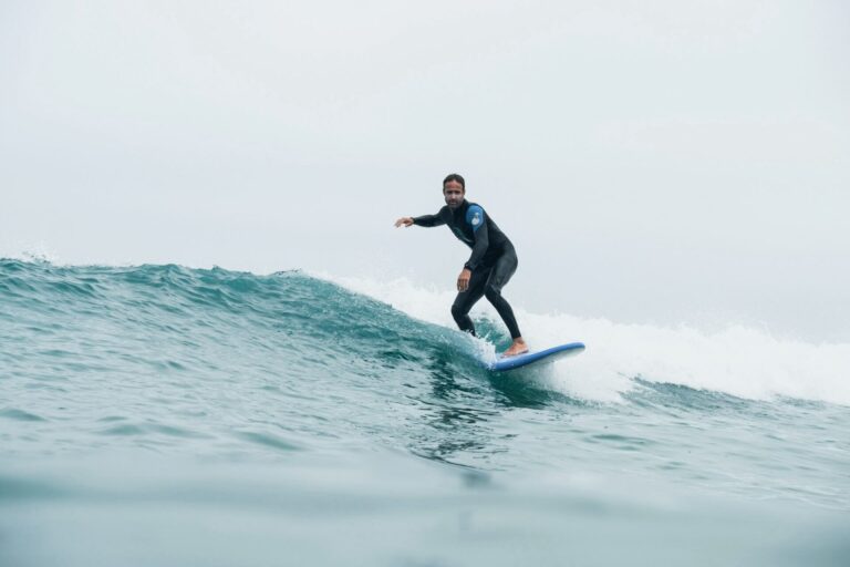 En person i en våddragt surfer på en lille bølge i havet. Det er en overskyet dag, og vandet er roligt. Surferen er balanceret i midten af bølgen, med deres arme strakt for stabilitet.