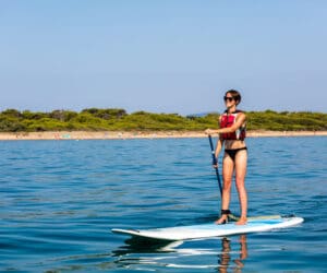 En person iført redningsvest og solbriller er på stand-up paddleboarding på en rolig vandmasse. Kystlinjen med træer og en sandstrand er synlig i baggrunden under en klar blå himmel.