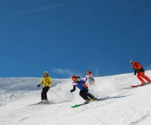 Fire skiløbere i farverige outfits glider ned ad en sneklædt skråning under en klar blå himmel. De er arrangeret i en forskudt formation, der hver viser forskellige skistile og skaber spor af sne bag dem.