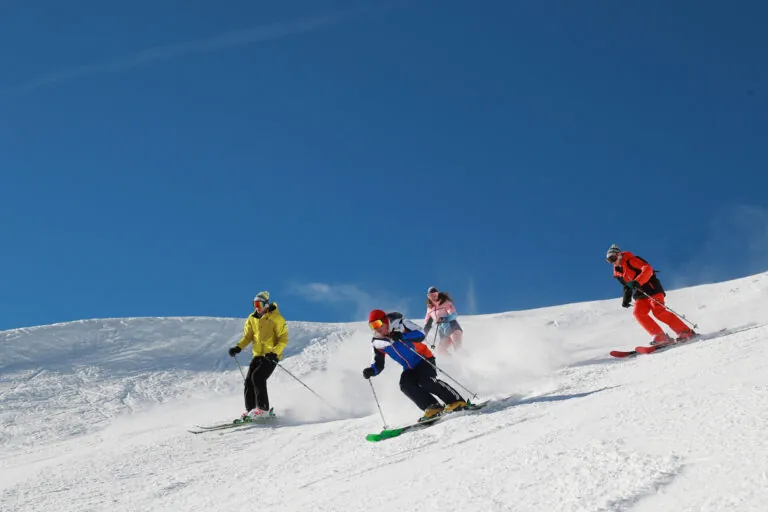 Fire skiløbere i farverige outfits glider ned ad en sneklædt skråning under en klar blå himmel. De er arrangeret i en forskudt formation, der hver viser forskellige skistile og skaber spor af sne bag dem.