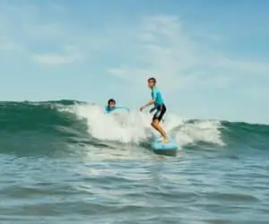 To unge surfere rider på en bølge under en klar blå himmel. De står begge på deres brædder og balancerer på vandet, mens bølgen krøller sig bag dem. Havet glimter i sollys og skaber en levende scene med surfing.