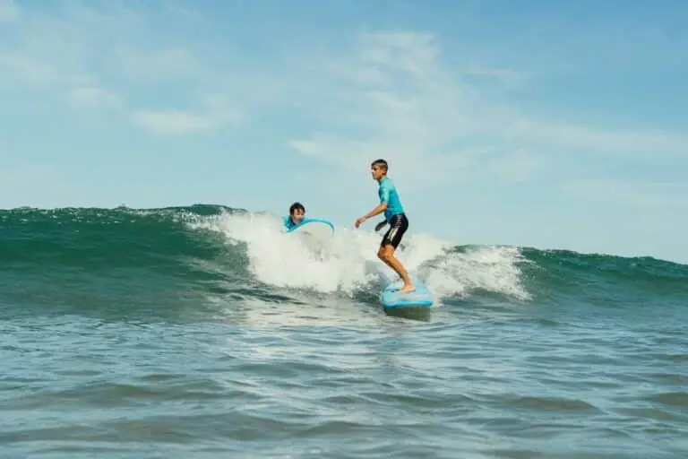 To unge surfere rider på en bølge under en klar blå himmel. De står begge på deres brædder og balancerer på vandet, mens bølgen krøller sig bag dem. Havet glimter i sollys og skaber en levende scene med surfing.