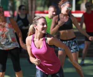 En gruppe mennesker, der deltager i en udendørs fitness- eller dansetime. Fokus er på en kvinde i en lyserød tanktop, der smiler og bevæger sig energisk. Andre i baggrunden er også engageret i aktiviteten, iklædt afslappet træningstøj.